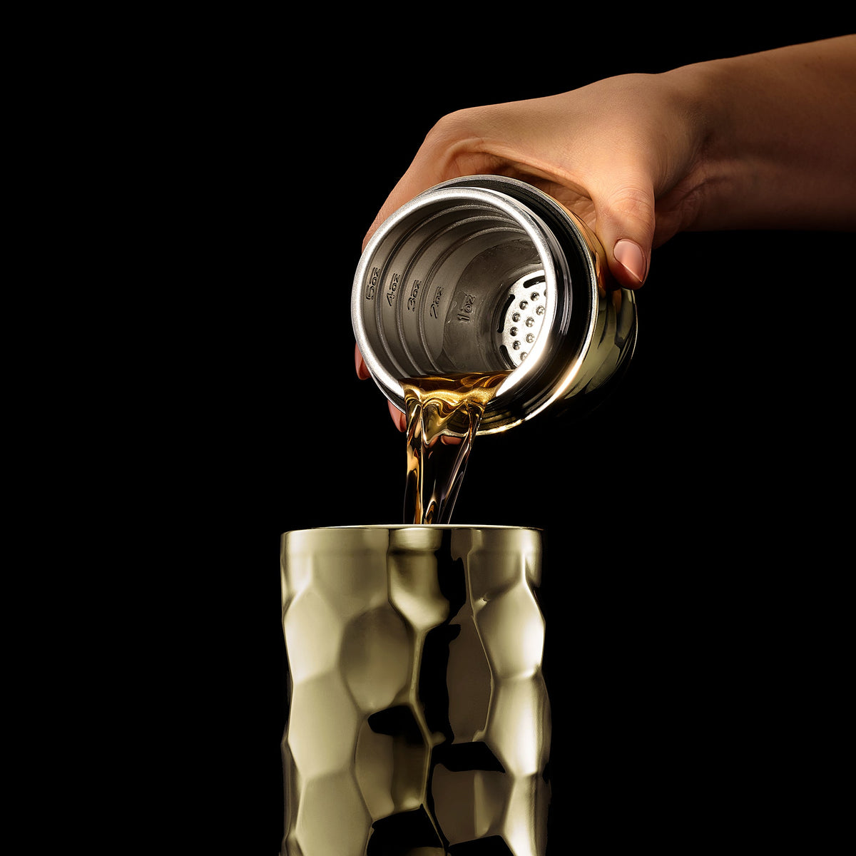 Hand pouring liquid from a hexclad cocktail shaker into another metallic container against a black background